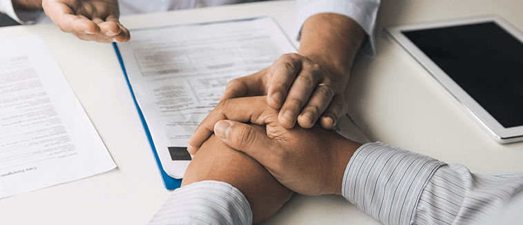 Two hands clasped together on a desk with documents and a tablet in the background, symbolizing collaboration and support during a consultation.