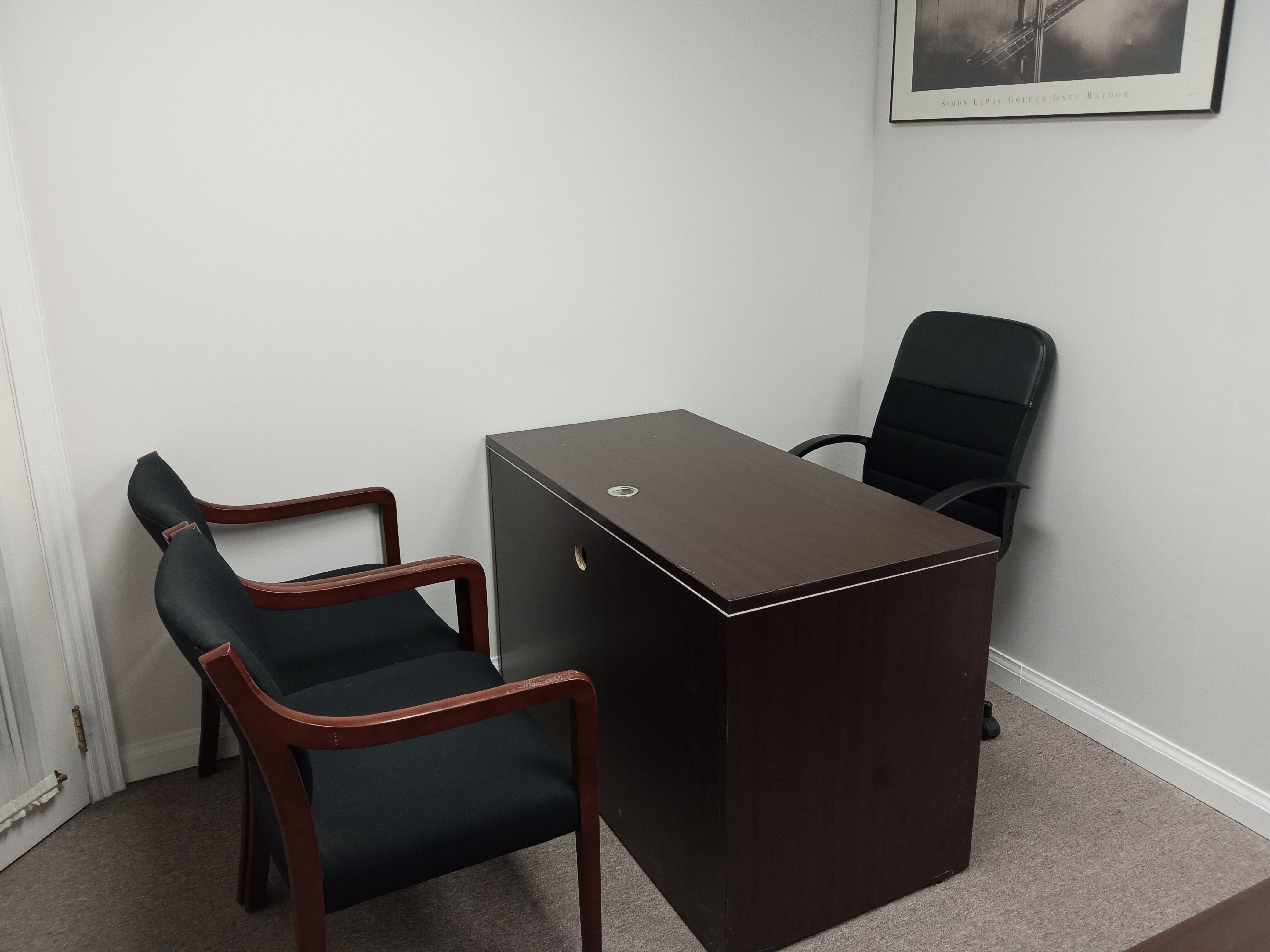 A small office space featuring a wooden desk and two black chairs, with a chair at the desk and a framed picture on the wall.
