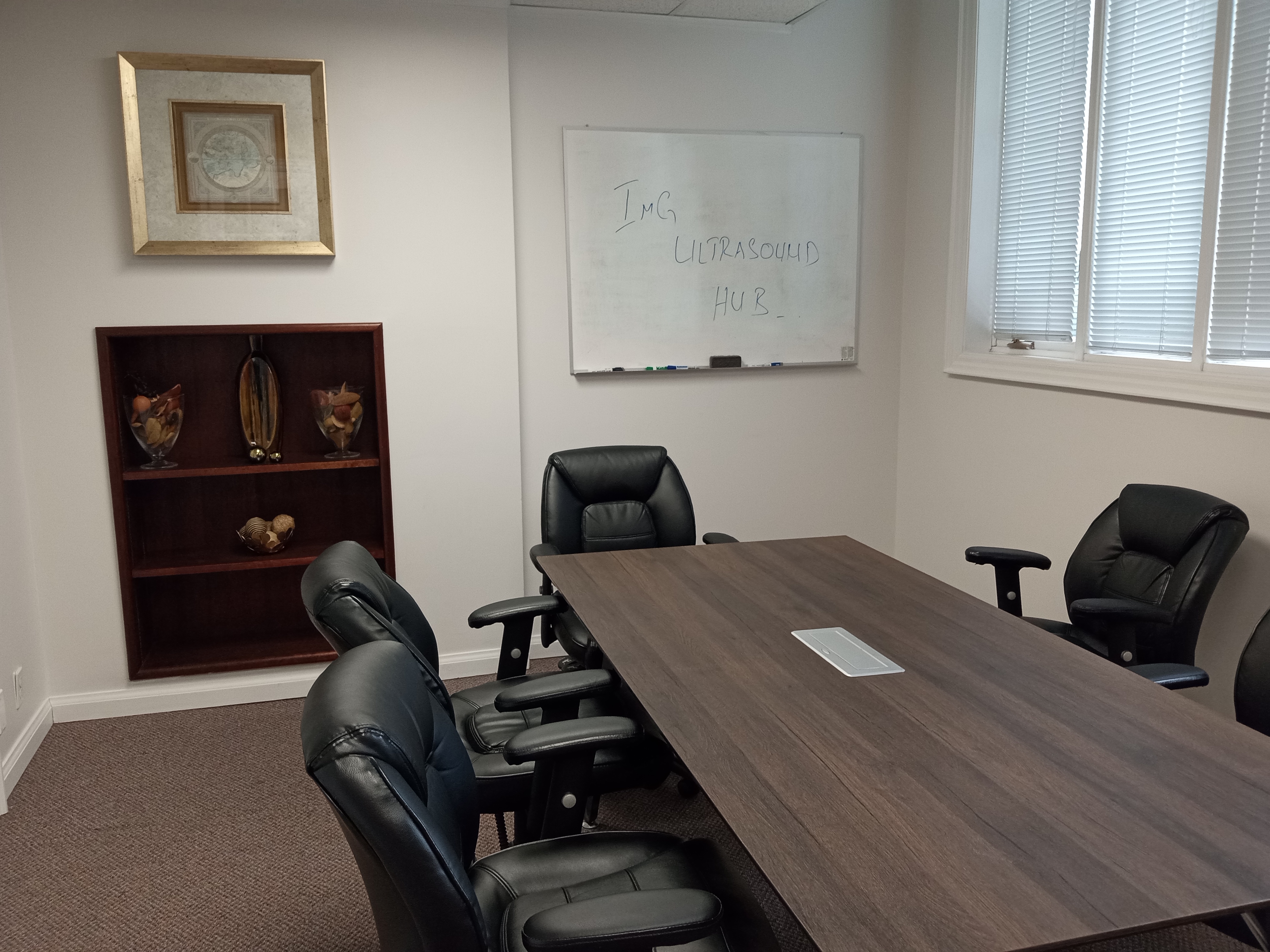 A meeting room at IMG Ultrasound Training Hub, featuring a large wooden table surrounded by black leather chairs, a whiteboard with 'IMG ULTRASOUND HUB' written on it, and a shelf displaying decorative vases.