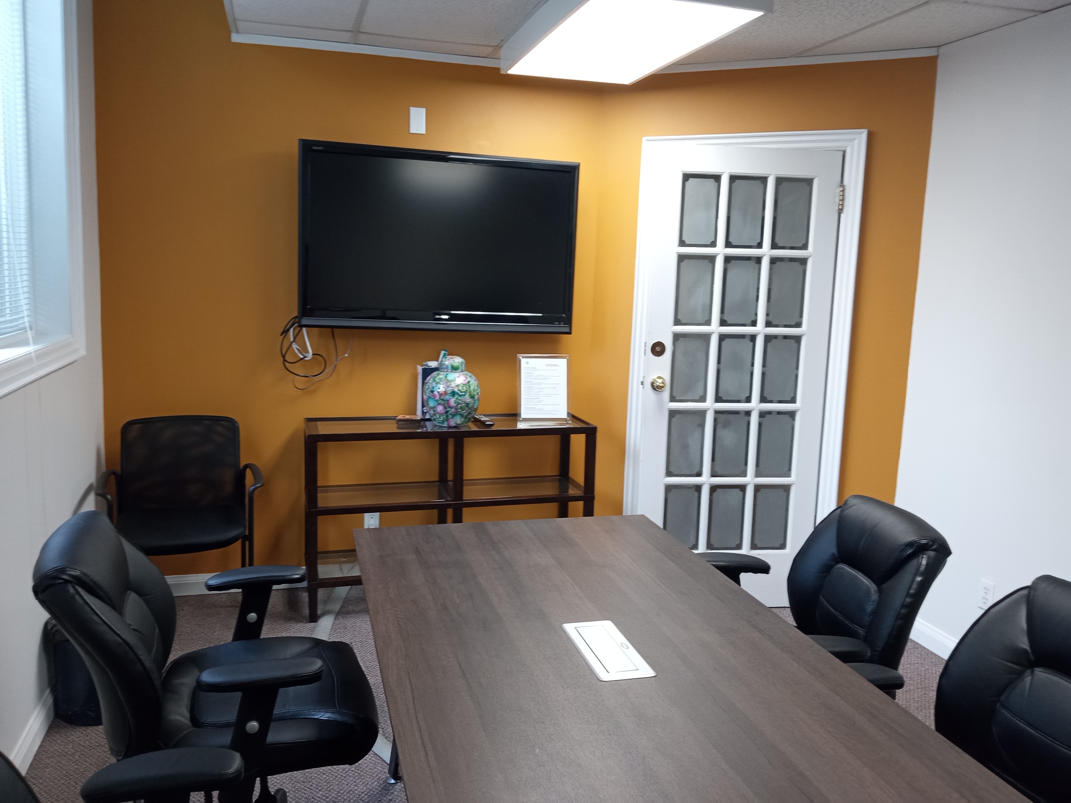 A meeting room with a wooden table, black chairs, and a large TV mounted on the wall, featuring a bright orange accent wall.
