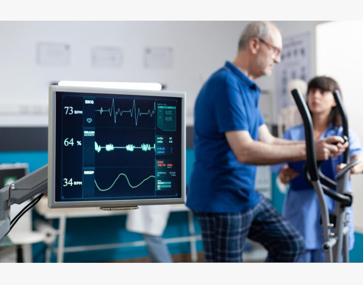 A monitor displaying vital signs including EKG, heart rate, and respiratory rate in a medical training setting, with a healthcare professional discussing with a patient.